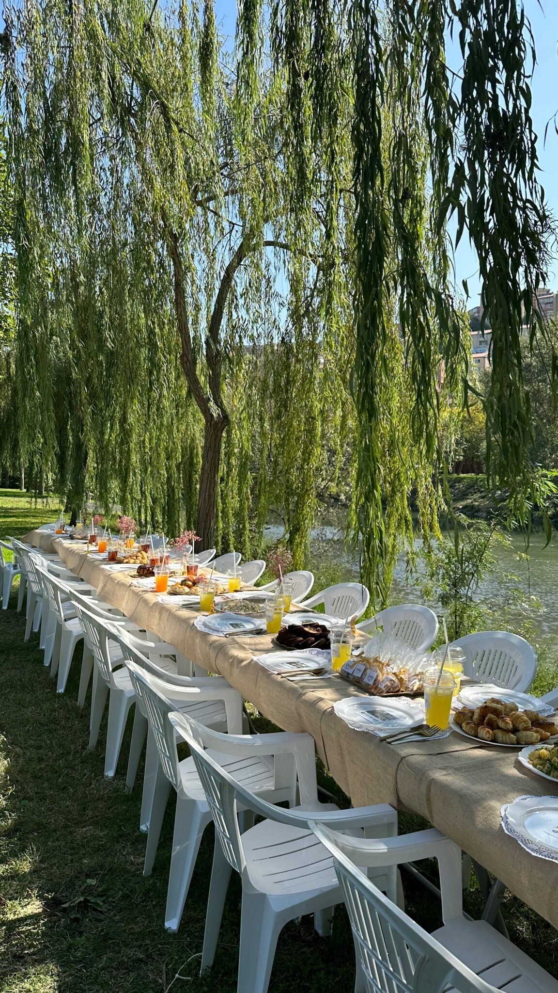Outdoor banquet setup with tables under willow trees by a peaceful river.